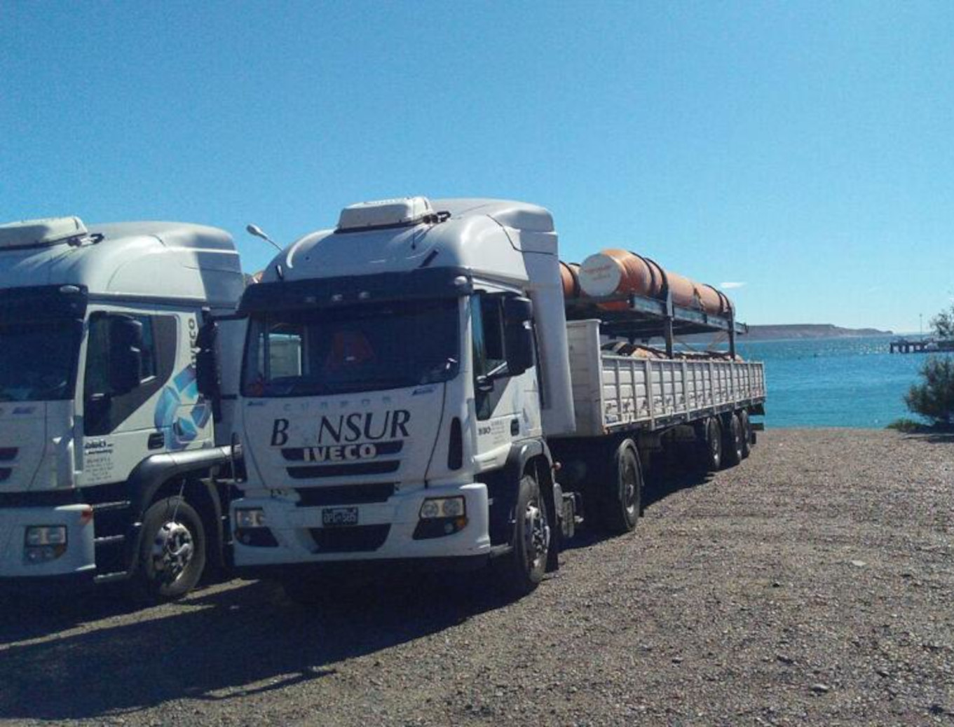 Camión blanco de la marca IVECO, de la empresa Bonsur, transportando carga cilíndrica, estacionado cerca de un cuerpo de agua