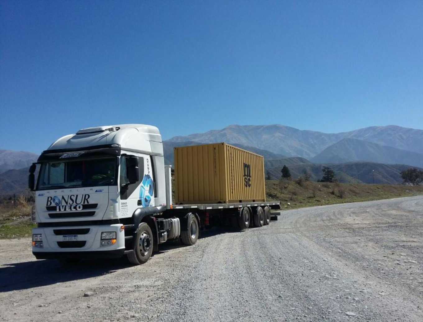 Camión blanco de la marca IVECO, de la empresa Bonsur, transportando un contenedor amarillo, en una ruta de montaña